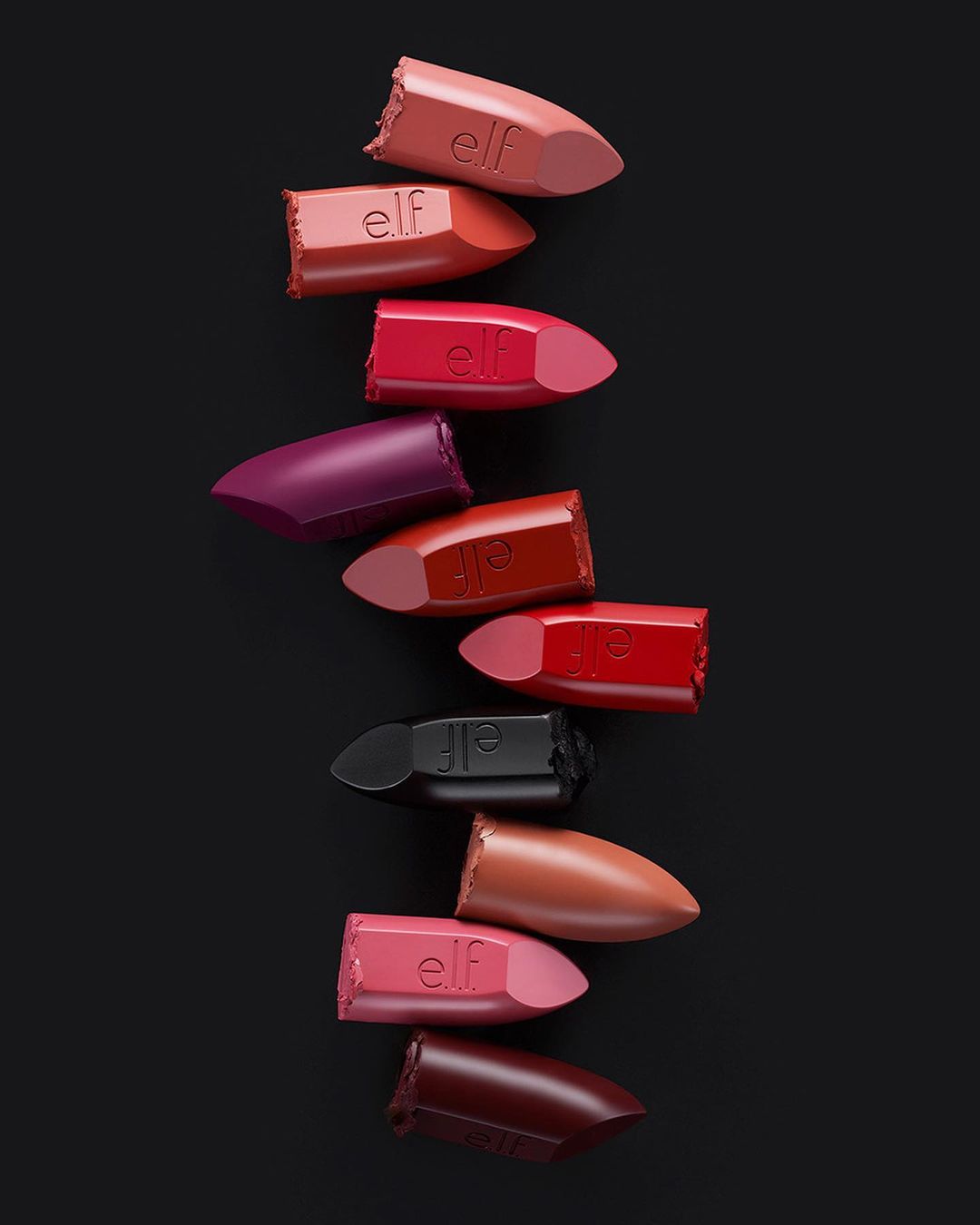Pile of e.l.f. lipsticks in various shades on a black background. Beauty still life of makeup products, arranged in a vertical composition.