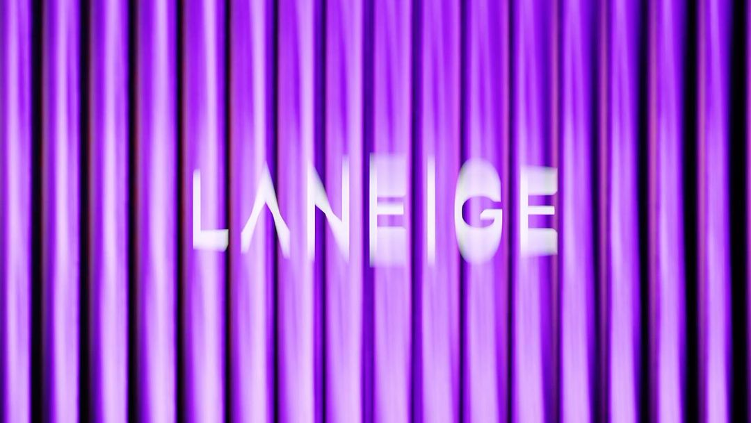 Close-up of the word "LANEIGE" in white font against a blurred, purple-toned curtain background, shot in a studio setting.