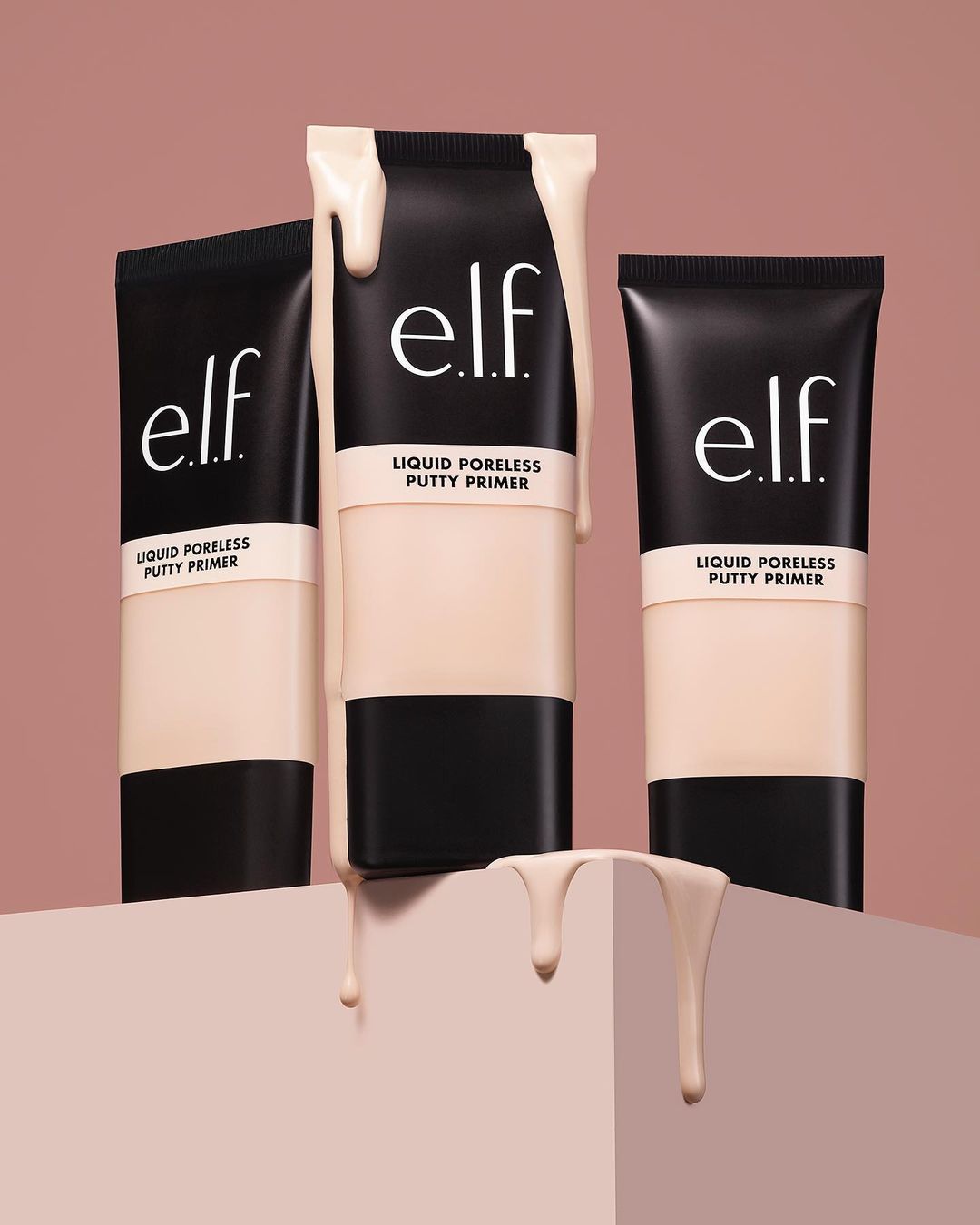 Three black tubes of e.l.f. Liquid Poreless Putty Primer are staged on a geometric stand against a light brown background, product spilling.