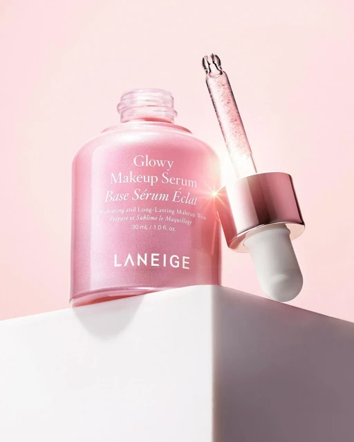 Close-up of a pink glass bottle of Laneige Glowy Makeup Serum on a white cube, with a dropper, against a gradient pink background.