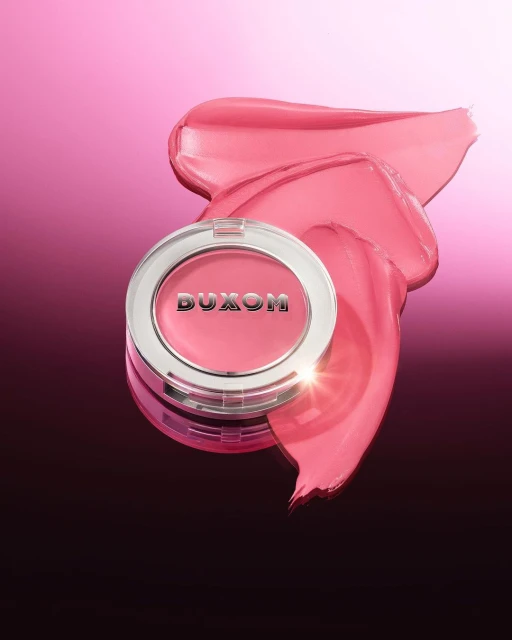 Buxom Cosmetics blush product in a silver container with a smear of matching pink-toned blush on a gradient pink background.