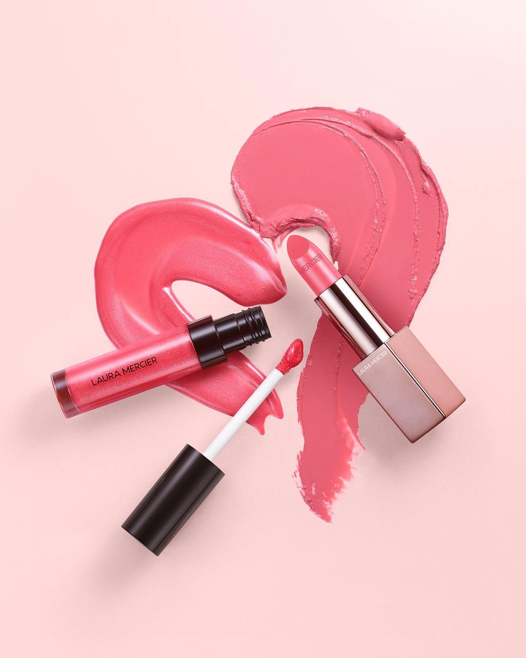 Cosmetic still life of lipstick and lip gloss with smears on a pink backdrop, advertising a beauty product in a studio setting.