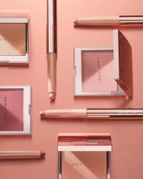 Top-down flat lay of Laura Mercier makeup products including blush compacts and eye shadow sticks on a pink background.