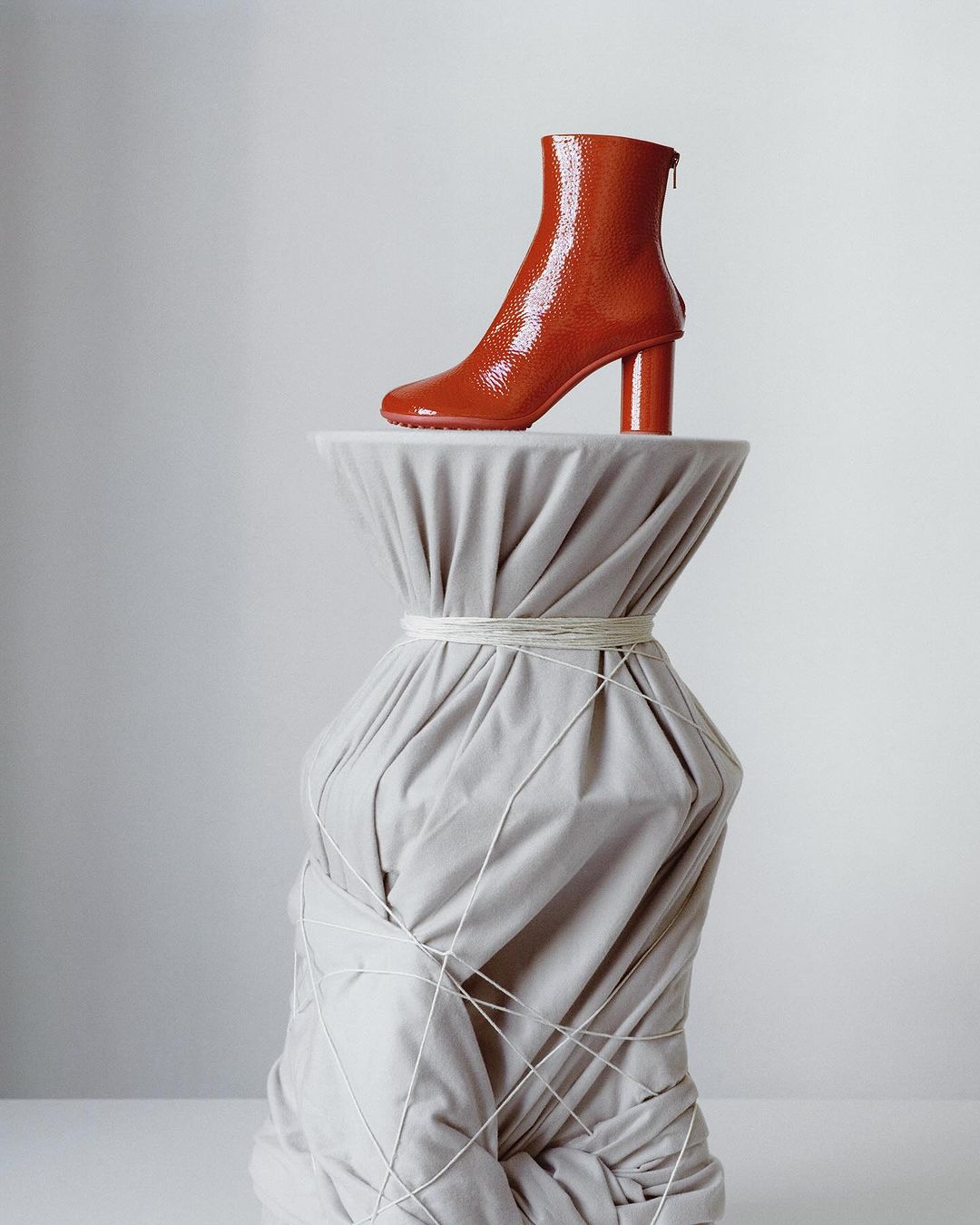 Studio shot of a shiny, red-brown, heeled ankle boot displayed on a draped pedestal against a neutral backdrop. Fashion still life.