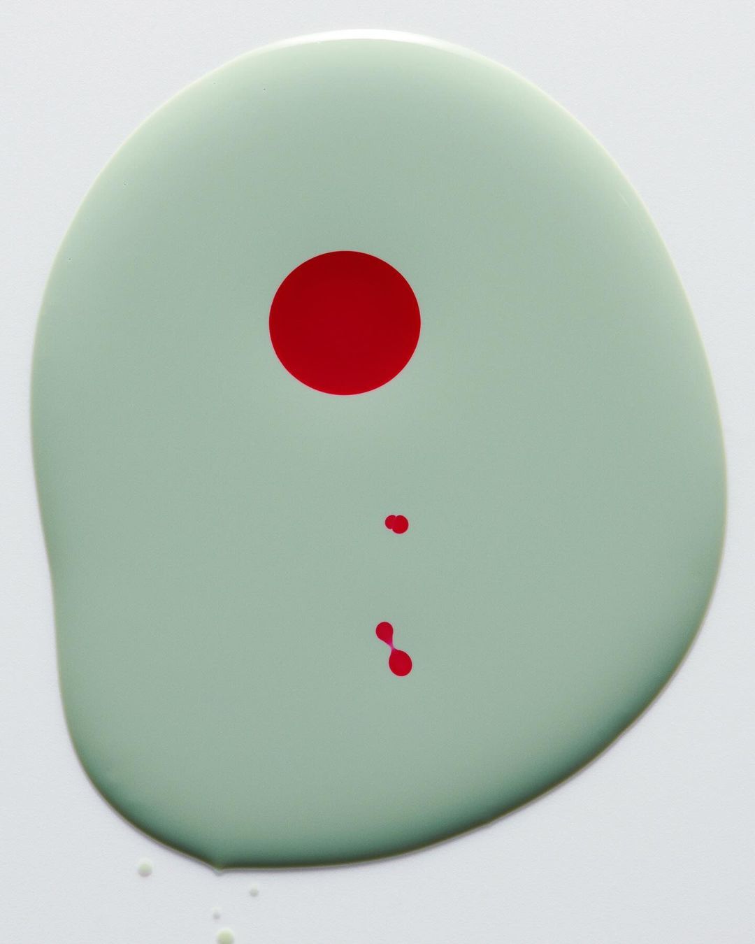 Overhead studio shot of a light green paint blob with a large red circle and two smaller droplets on a white surface.