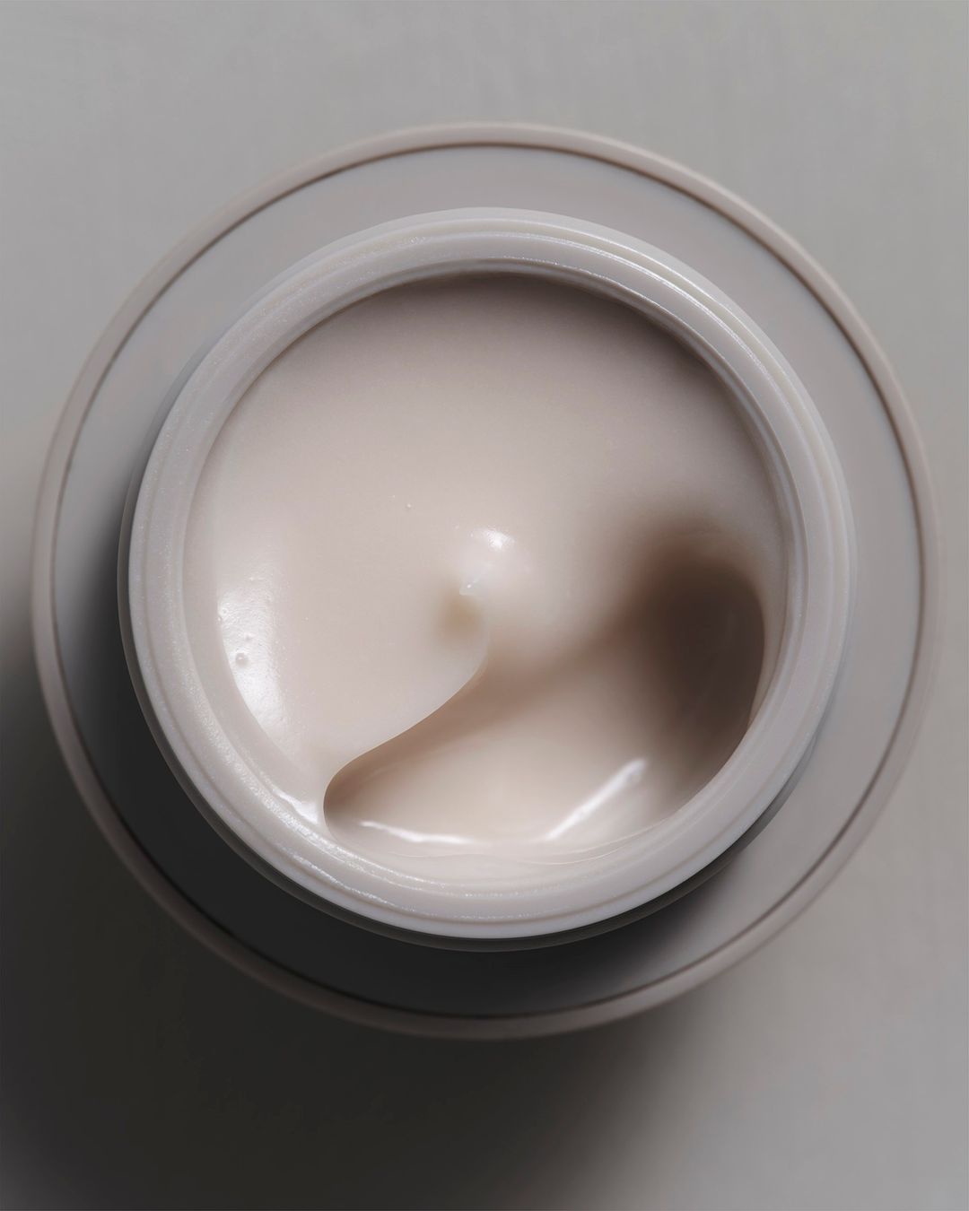Close-up, overhead shot of skincare cream in a grey jar. The texture of the cream is smooth and light beige, studio lighting.