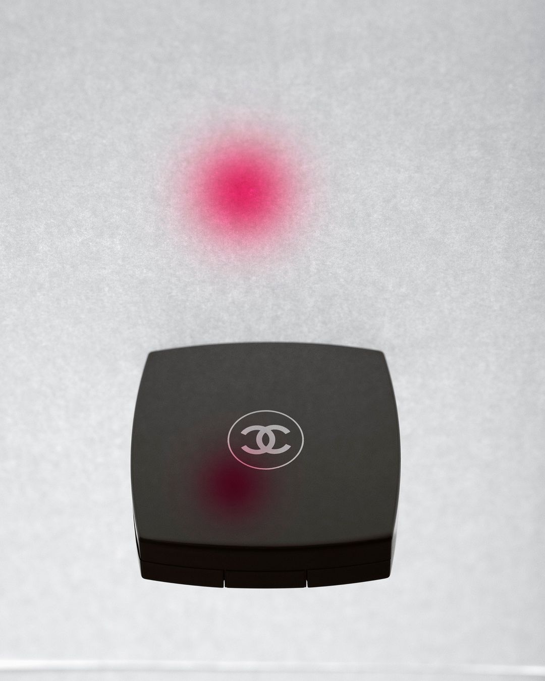 Close-up of a black Chanel compact against a textured white background with a pink, soft spot light shining on the compact's logo.