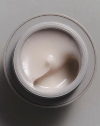 Top-down view of an open jar of pale cream cosmetic product with a smooth, swirled surface against a neutral grey background.