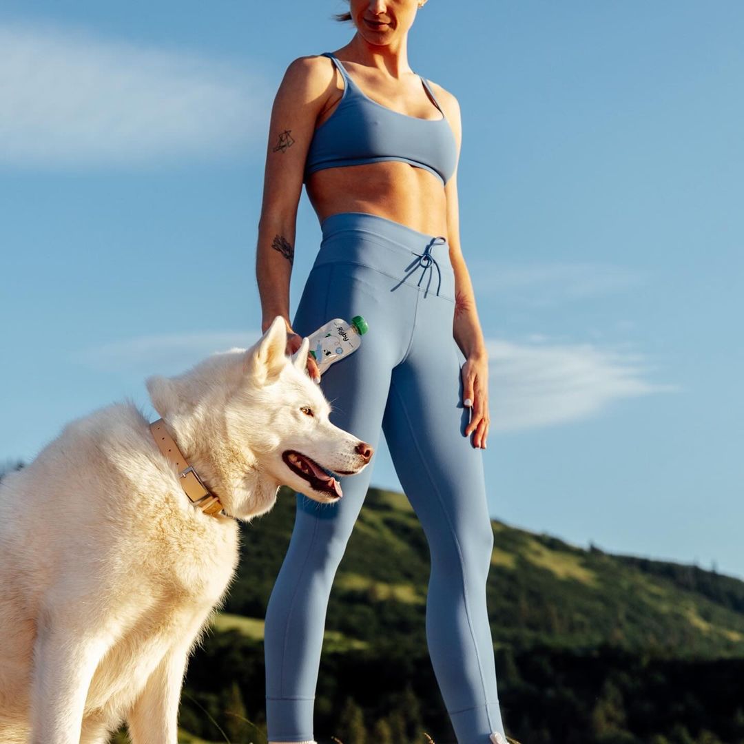 A slender adult woman in athletic wear stands with a white dog outdoors against a sunny sky and distant mountains, post-workout hydration.
