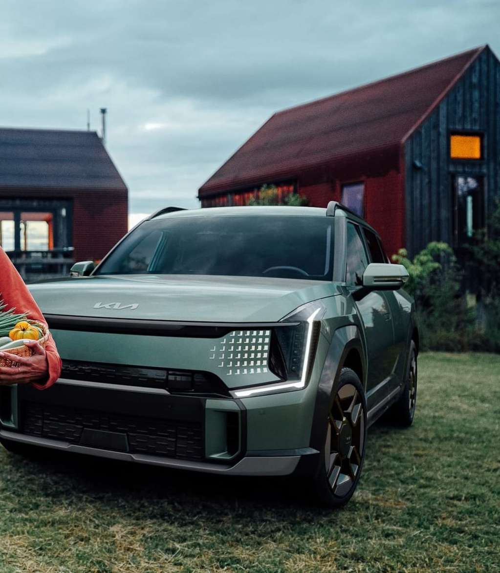 A light-green Kia SUV stands on a lawn in front of rural buildings, with a person holding a basket of vegetables to the left.