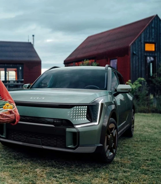 A light-green Kia SUV stands on a lawn in front of rural buildings, with a person holding a basket of vegetables to the left.