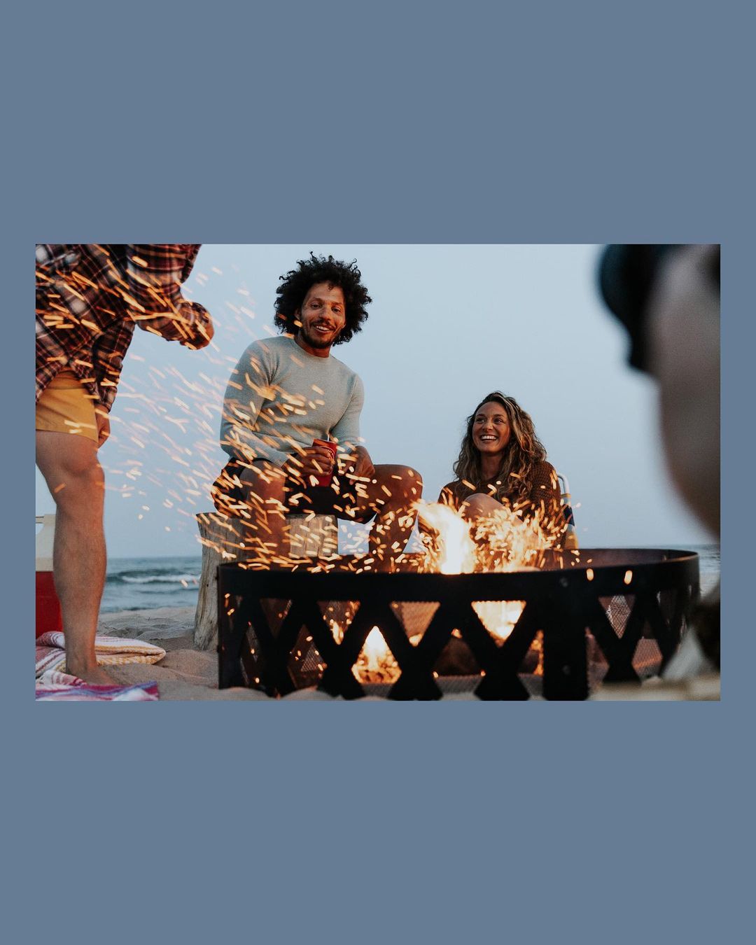 Three people sit around a fire pit on a sandy beach at dusk, two of them smiling, with the ocean and sky visible in the background.