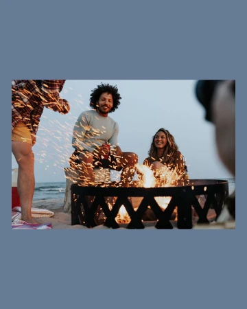 Three people sit around a fire pit on a sandy beach at dusk, two of them smiling, with the ocean and sky visible in the background.