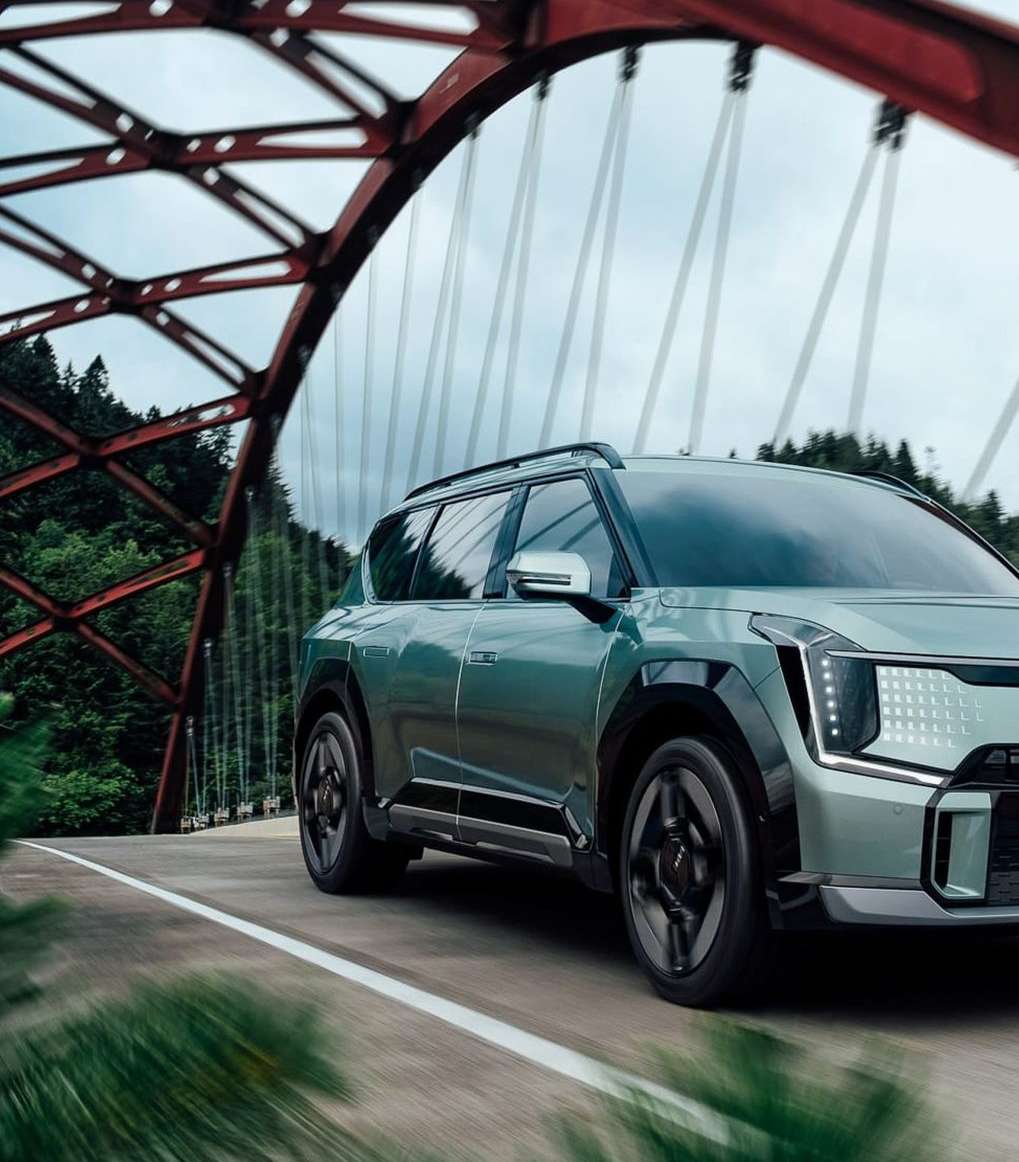 An SUV crosses a vintage red metal bridge with a forest backdrop on an overcast day, showcasing automotive design and scenic travel.