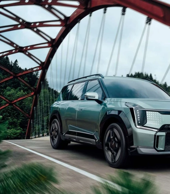 An SUV crosses a vintage red metal bridge with a forest backdrop on an overcast day, showcasing automotive design and scenic travel.