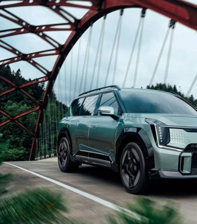An SUV crosses a vintage red metal bridge with a forest backdrop on an overcast day, showcasing automotive design and scenic travel.