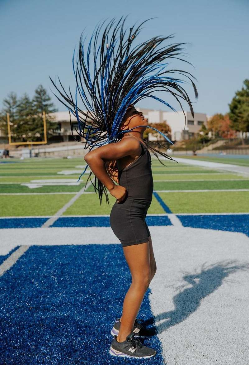 An African-American woman in black sportswear stands on a blue-and-green field with her arms on her hips and feathered headwear.