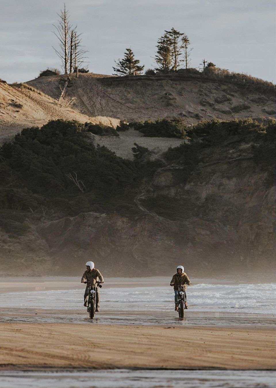 Two people riding electric bikes on a beach with a dune and trees in the background on a hazy day.