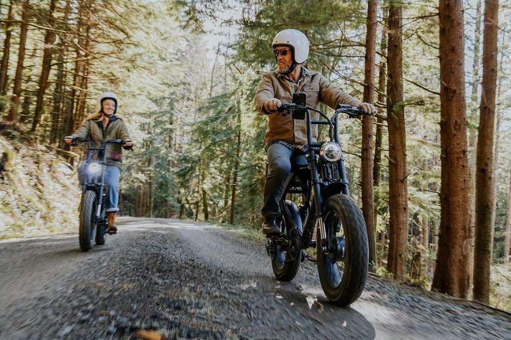Two people riding electric bikes on a dirt path through a sun-dappled forest, wearing helmets and casual clothing, enjoying an outdoor activity.