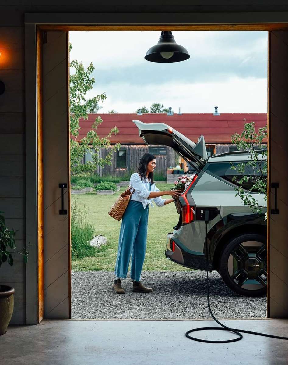 Woman loads flowers into an electric vehicle (EV) parked outside a barn. The EV is plugged in for charging in a rural setting.