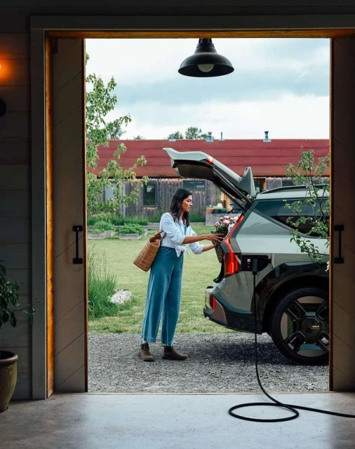 Woman loads flowers into an electric vehicle (EV) parked outside a barn. The EV is plugged in for charging in a rural setting.