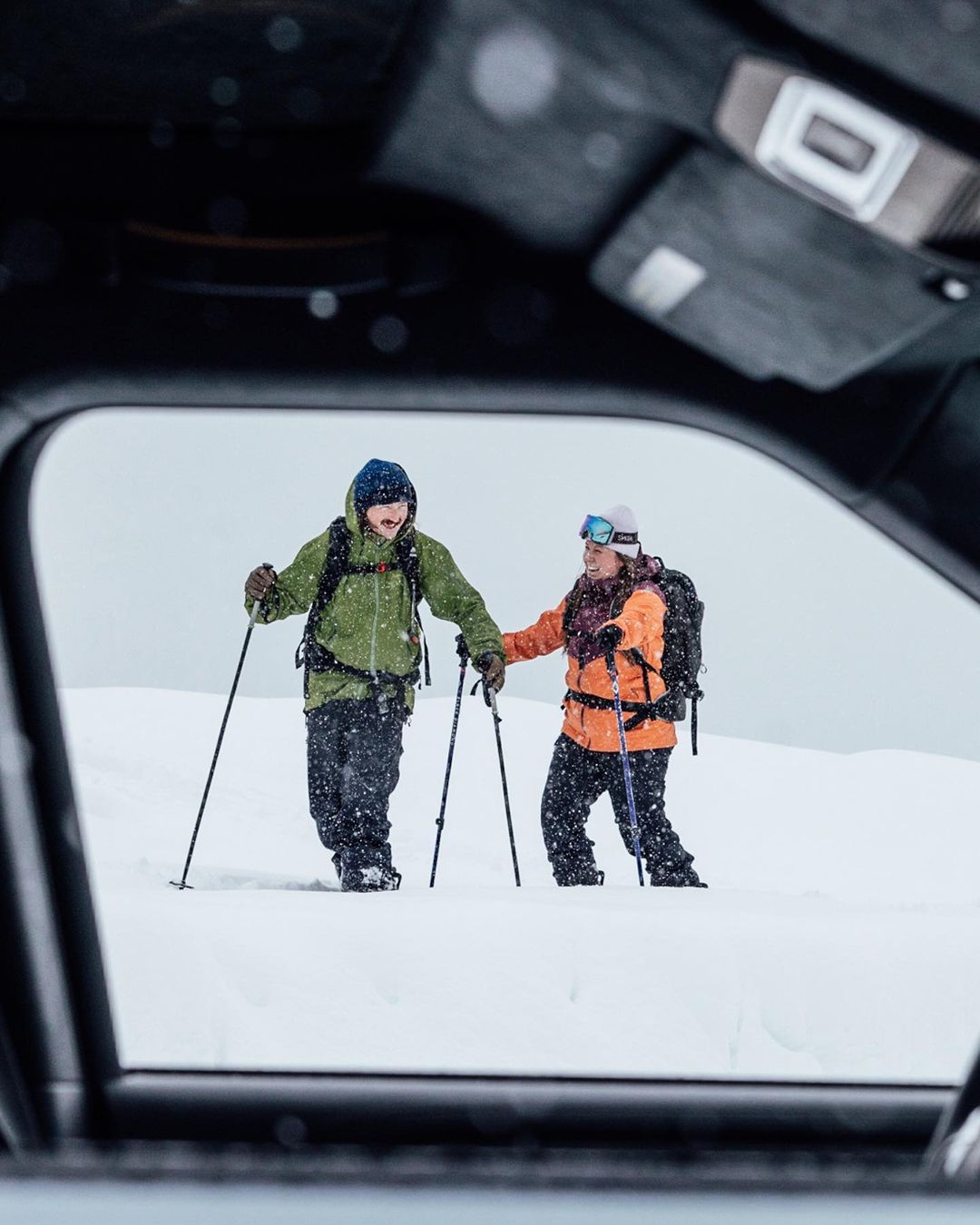 Two people snowshoeing uphill on a snowy landscape, viewed through a car window, wearing winter clothing, holding trekking poles.