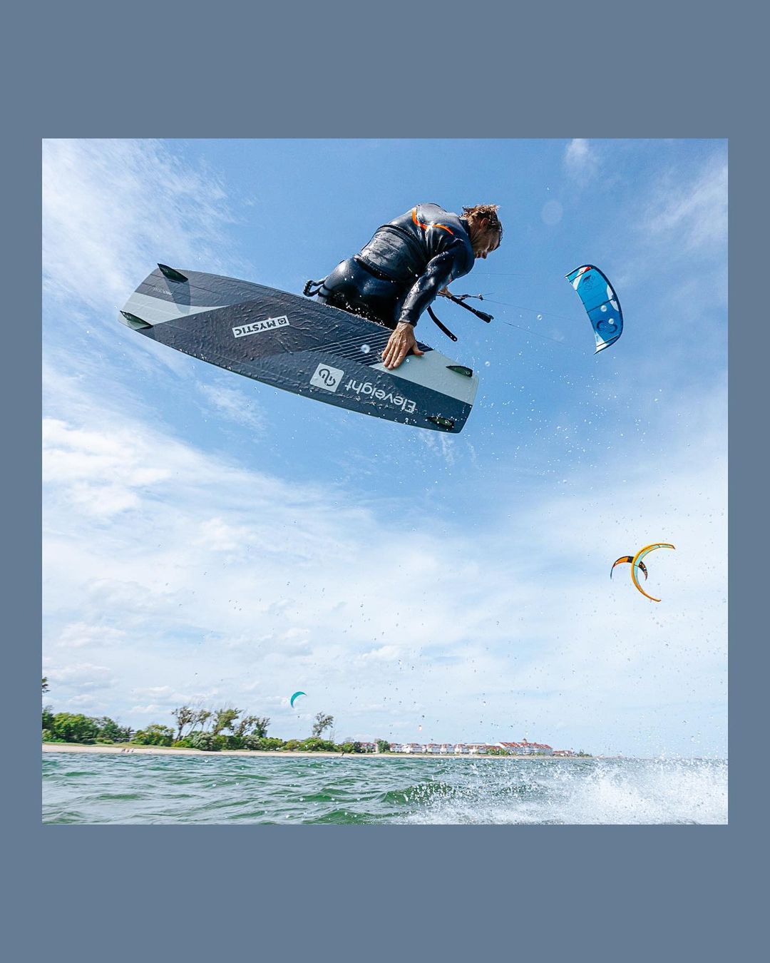 A kiteboarder in a black wetsuit soars through the air on a board with a kite in the background against a blue sky with some clouds.