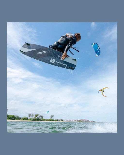 A kiteboarder in a black wetsuit soars through the air on a board with a kite in the background against a blue sky with some clouds.