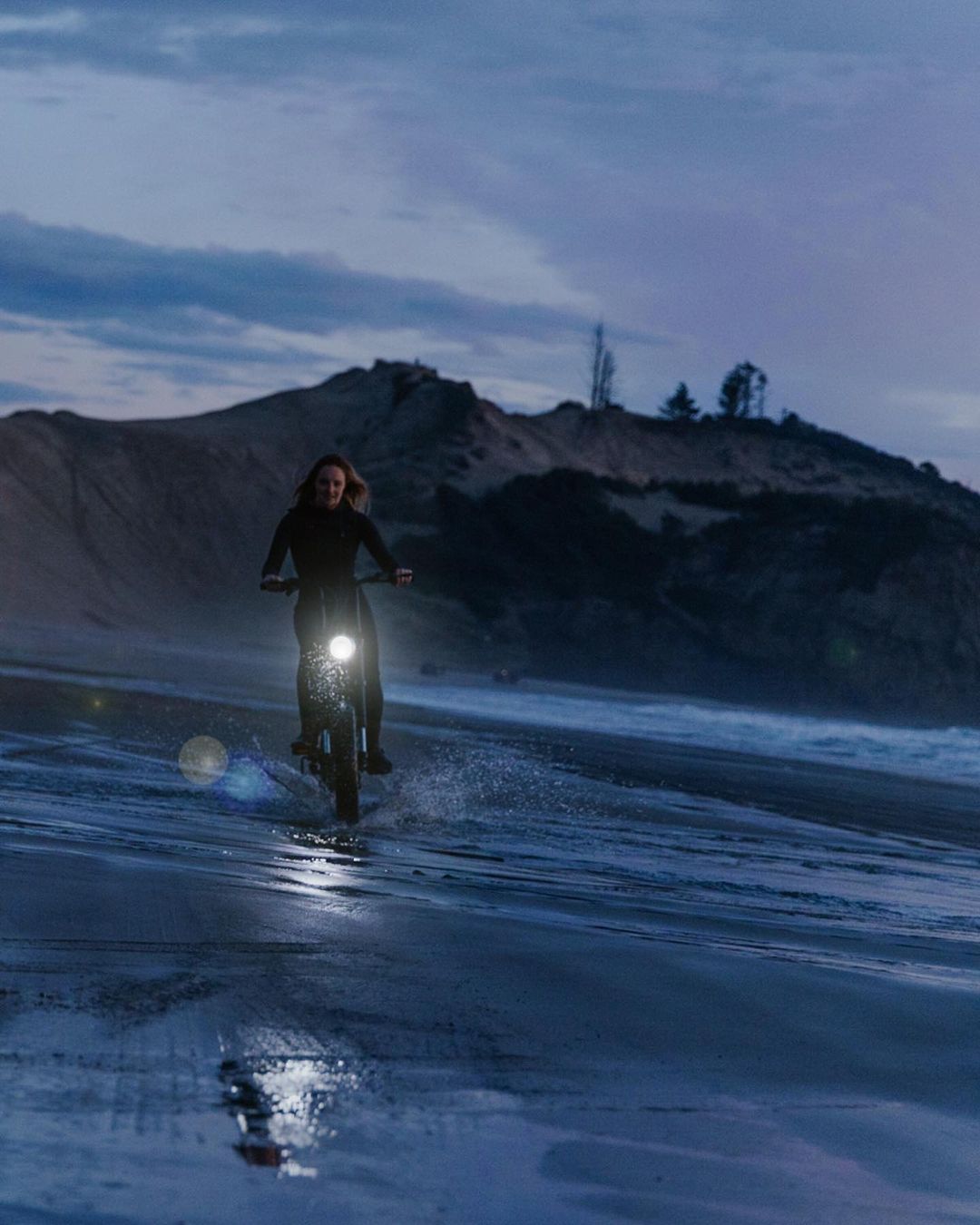 Woman rides an electric motorbike on a wet sandy beach at dusk, headlamp on, with a dark blue cloudy sky and a sandy dune background.