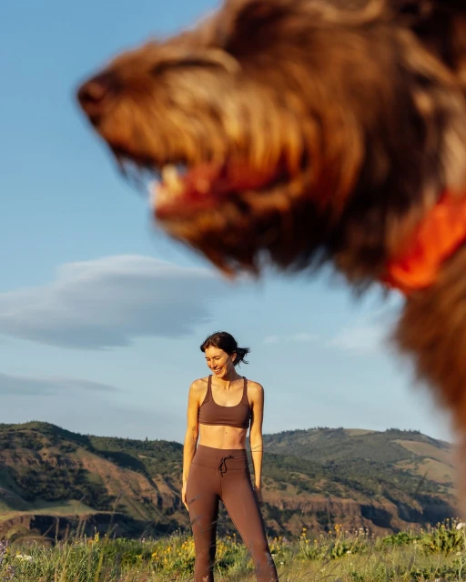 Woman in sportswear smiles in an outdoor meadow, with rolling hills and sky in the background; out-of-focus dog in the foreground.