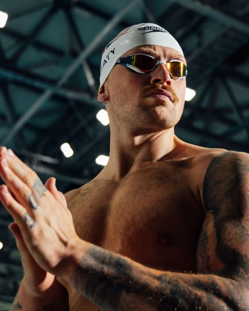 Close-up of a muscular adult male swimmer with a swim cap, goggles, and tattoos, clapping his hands indoors, low angle, looking right.
