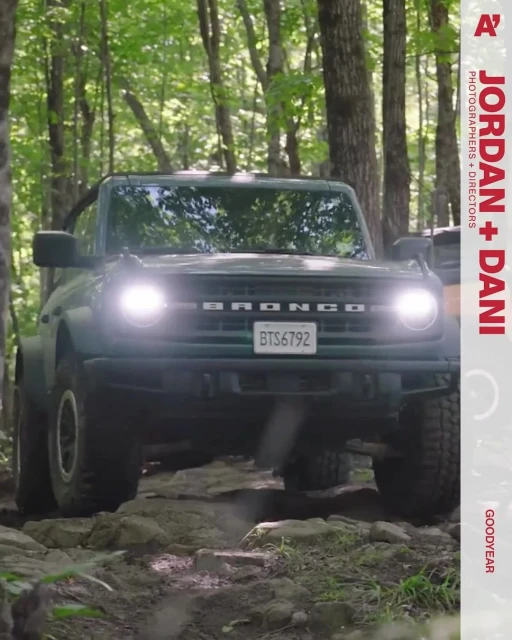 Green Ford Bronco four-wheel-drive car on a rocky path through a forest. The vehicle's headlights are on, with trees behind it.