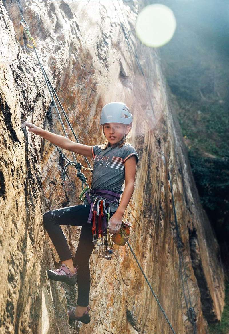 Young female rock climber in a helmet scales a textured rock wall with ropes and climbing gear on a sunny day outdoors.