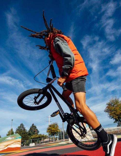 A young adult male performs a BMX bike trick in mid-air against a bright blue sky at a park. He wears casual clothing and Vans socks.