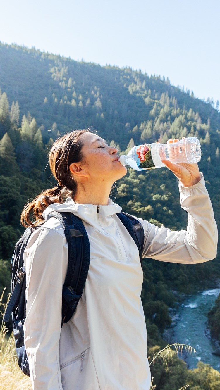 Woman in a white jacket with a dark backpack drinking from a plastic water bottle outdoors with a forested mountain and river background