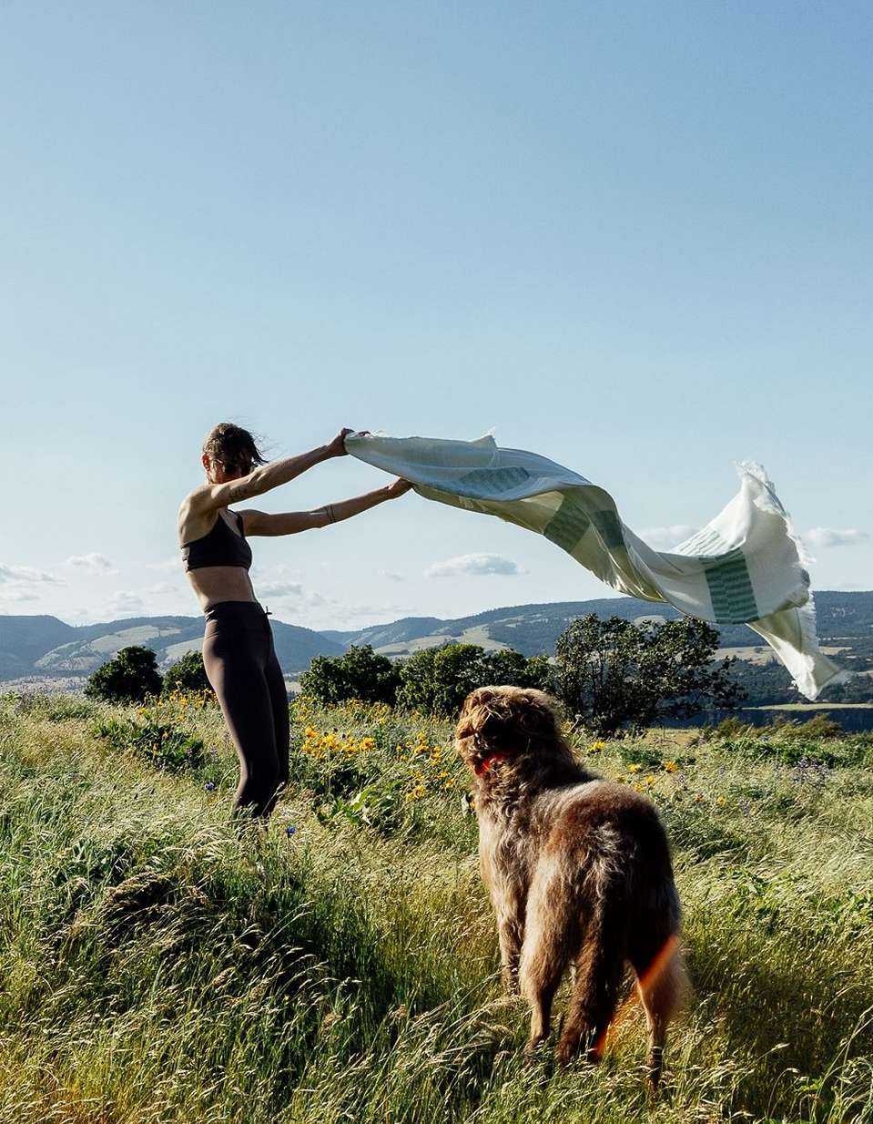 Woman in dark activewear holding a large patterned towel blowing in the wind in a grassy field with a dog in the foreground