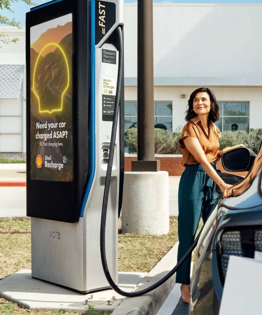Adult woman charges an electric car at a Volta charging station outdoors on a sunny day, with a Shell Recharge advertisement displayed.