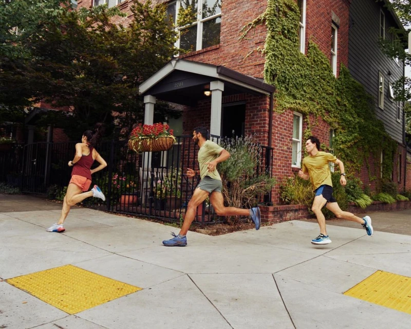Three people run along a sidewalk in an urban neighbourhood on a sunny day. Red brick buildings with green vines are visible.