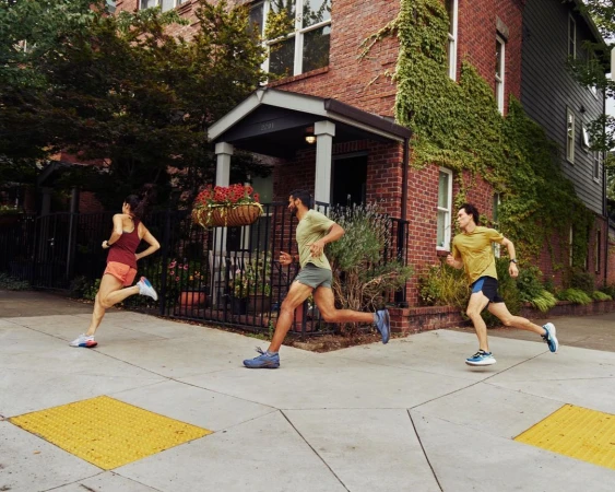 Three people run along a sidewalk in an urban neighbourhood on a sunny day. Red brick buildings with green vines are visible.