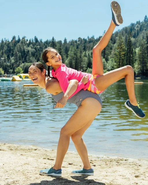 A woman gives a girl a piggyback ride on a sandy beach near a lake with lush green trees in the background, creating a joyful summer scene.