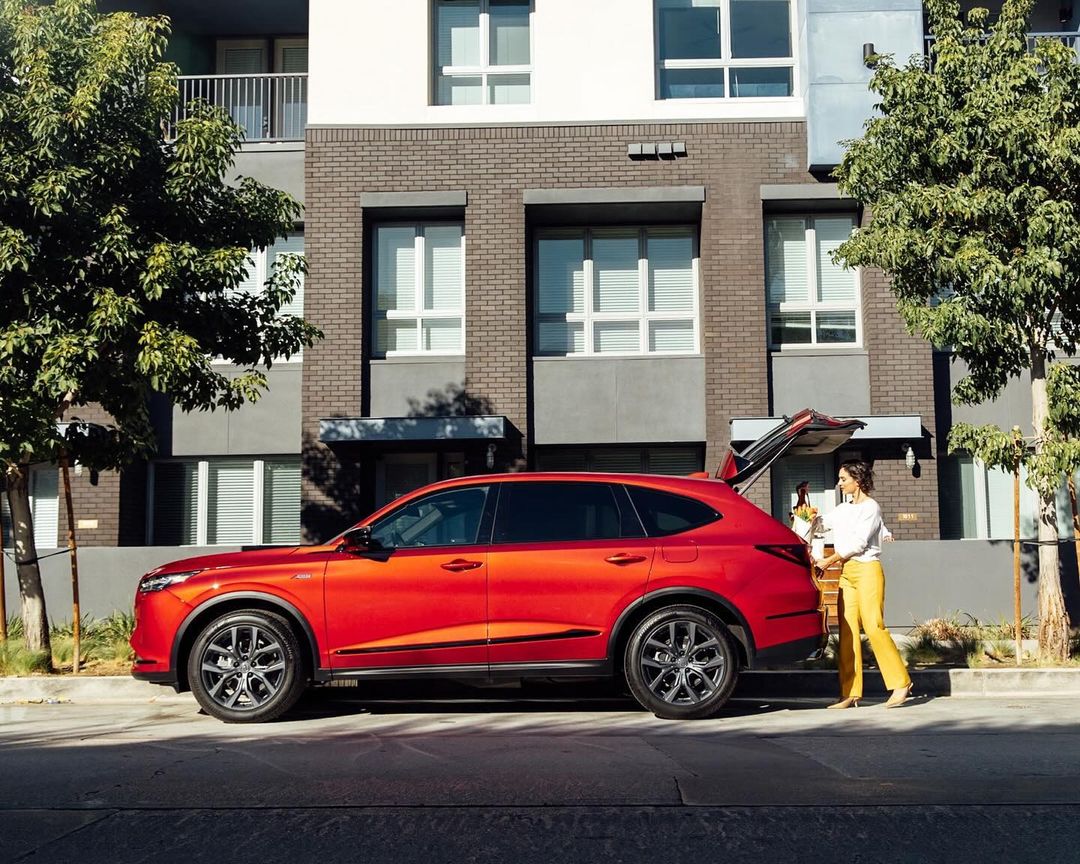 An orange SUV parked on a city street, a woman placing a basket in the open trunk, modern apartments in the background.