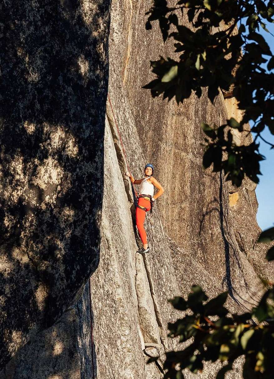 A woman in a climbing helmet and climbing harness scales a tall, rocky cliff face on a sunny day, using ropes for safety.