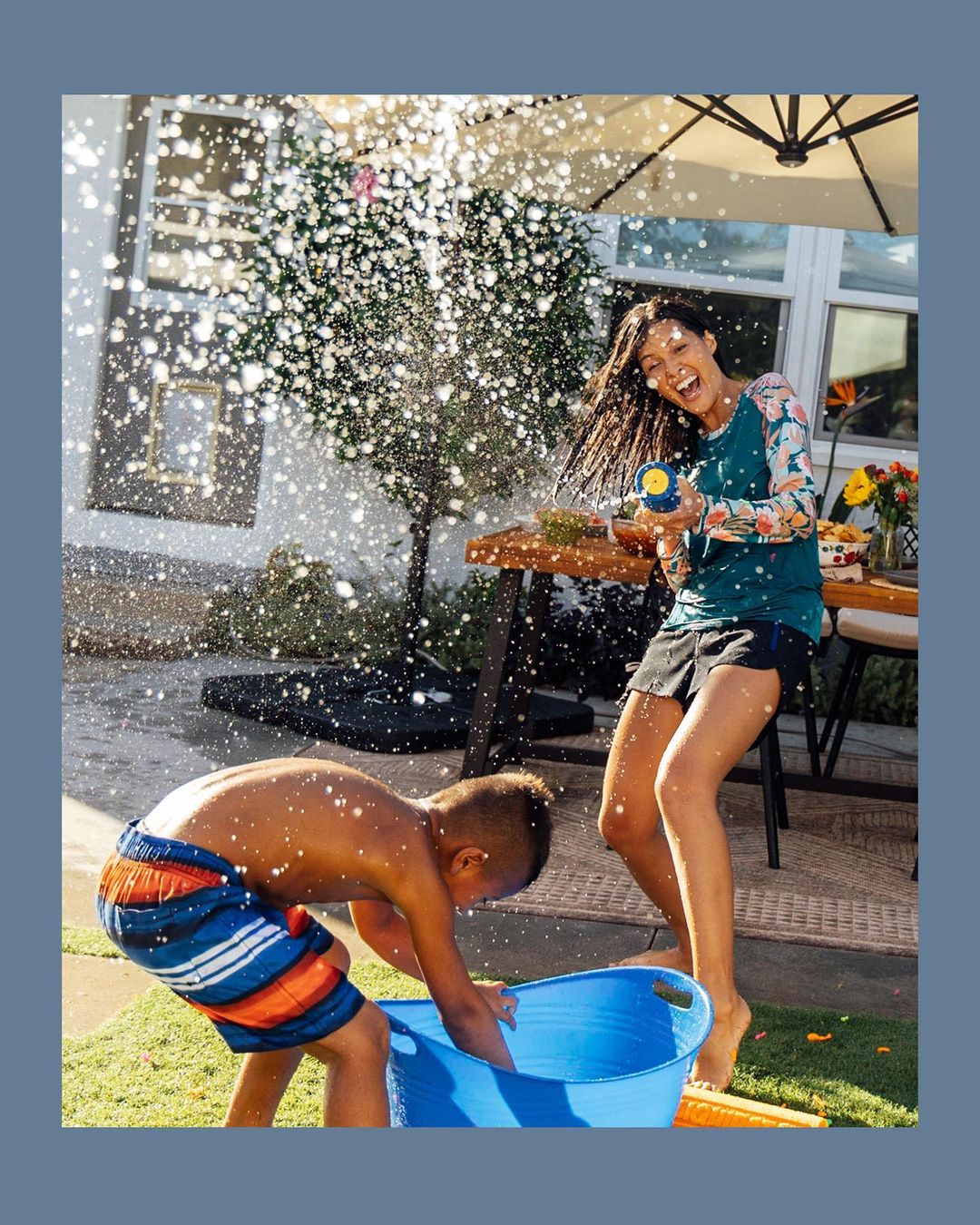 A woman and child play with water outside a house; the woman aims a water gun and the child holds a bucket under a shower of water.