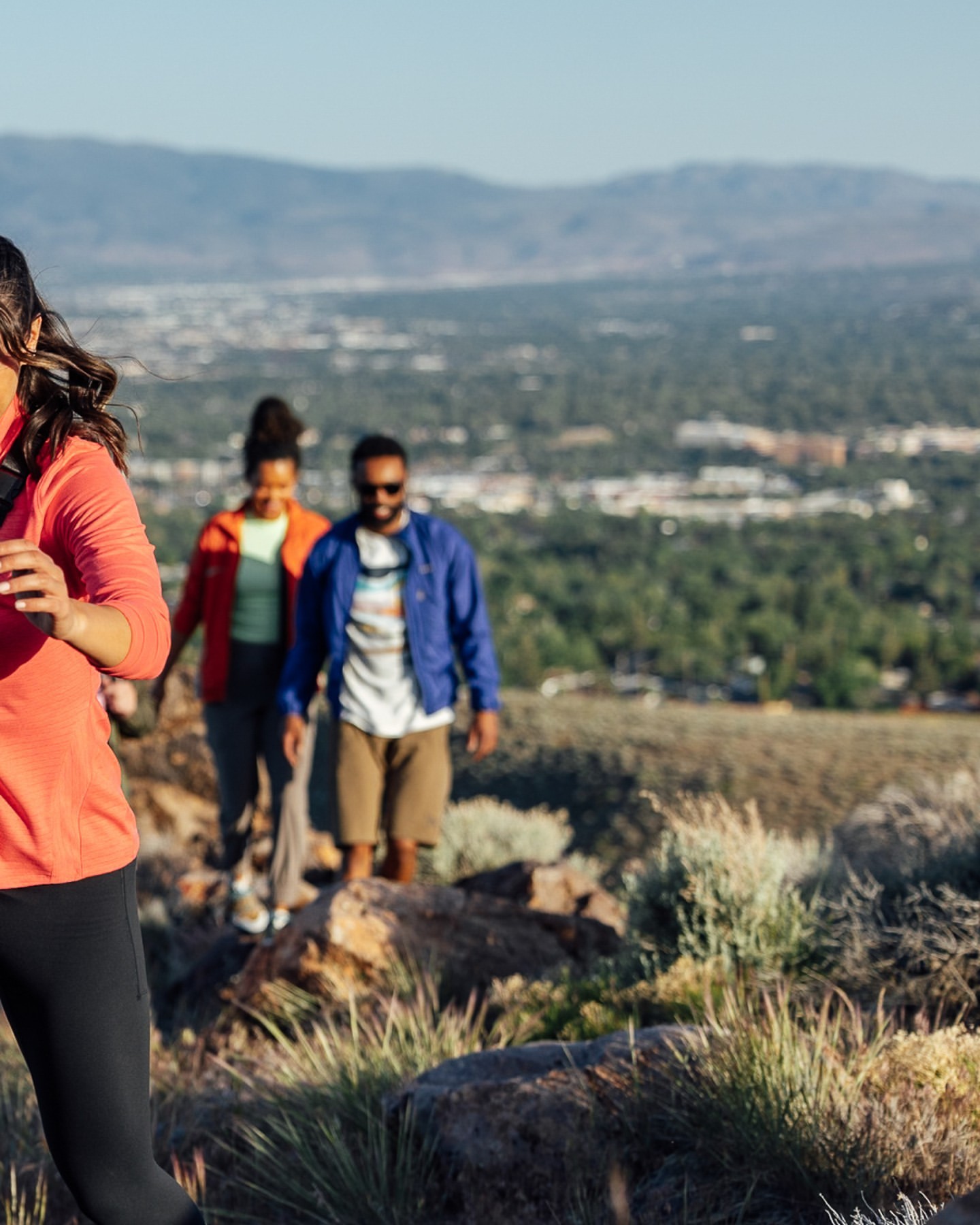 Three hikers on a mountain trail. A woman in coral leads a woman in orange and a man in blue. A city lies in the distance.
