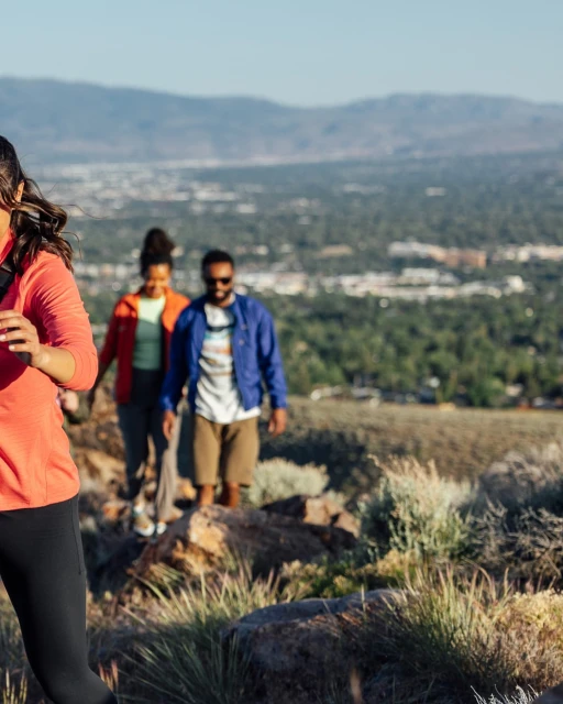 Three hikers on a mountain trail. A woman in coral leads a woman in orange and a man in blue. A city lies in the distance.