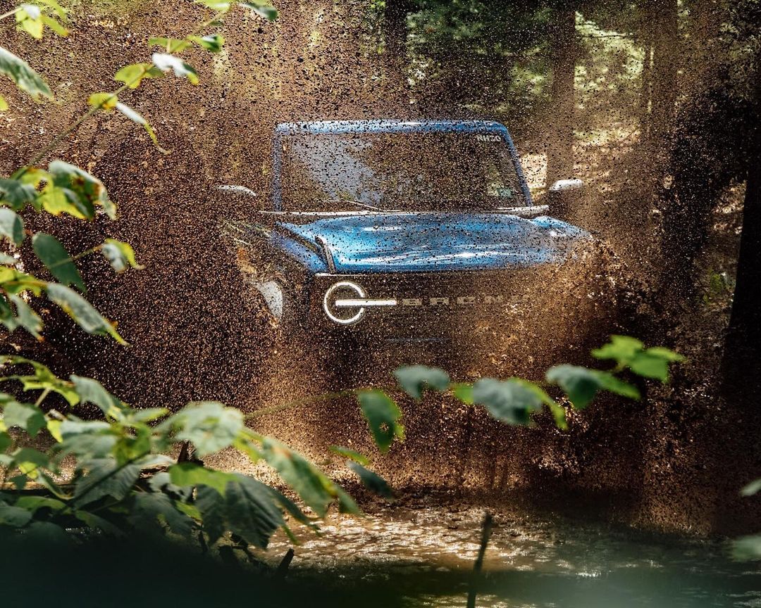Blue Ford Bronco splashes through a muddy stream in a forest, showcasing its off-road capabilities with natural green foliage.