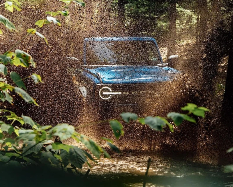Blue Ford Bronco splashes through a muddy stream in a forest, showcasing its off-road capabilities with natural green foliage.
