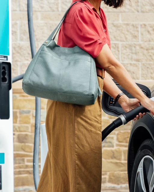 Woman charges electric car with a green leather shoulder bag in a stylish outdoor setting with a charging station.