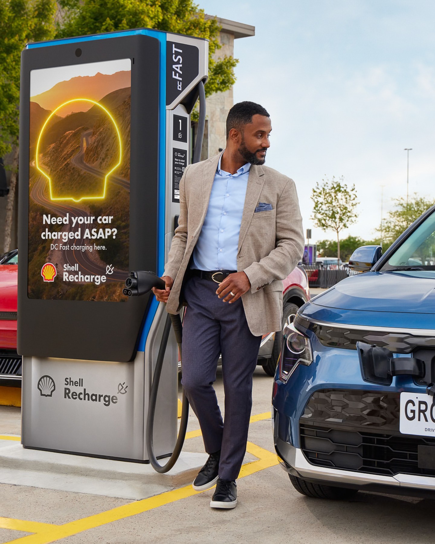Man connects charging cable to a light blue electric vehicle at a Shell Recharge station on a sunny day, promoting EV charging.