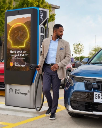 Man connects charging cable to a light blue electric vehicle at a Shell Recharge station on a sunny day, promoting EV charging.